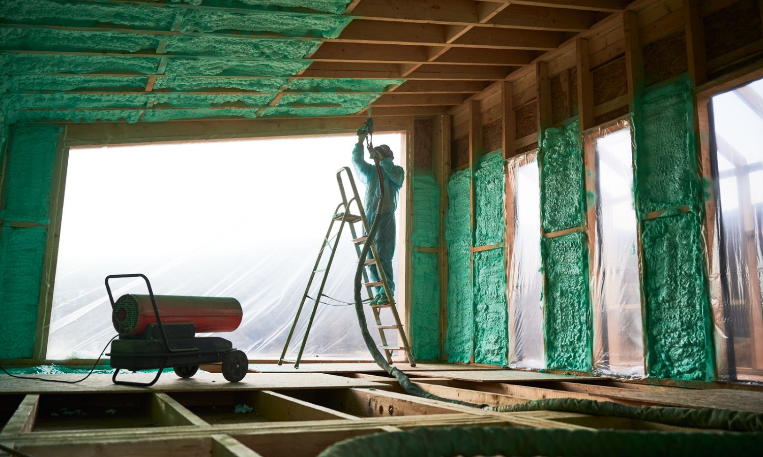 A man stands on a ladder, working in a room filled with spray foam insulation materials, focused on his task