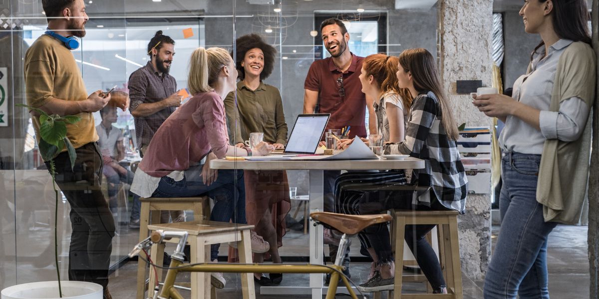 Group of coworkers smiling and chatting around a table in a modern office setting.