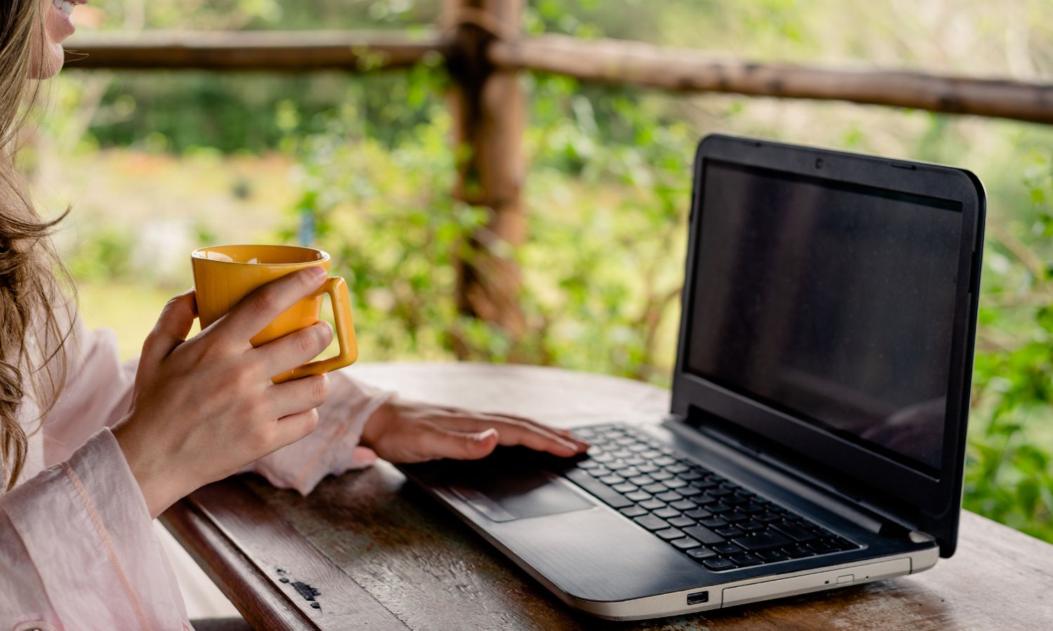 Person holding a yellow mug while working on a laptop with Microsoft SharePoint outdoors with a wooden table and greenery in the background.